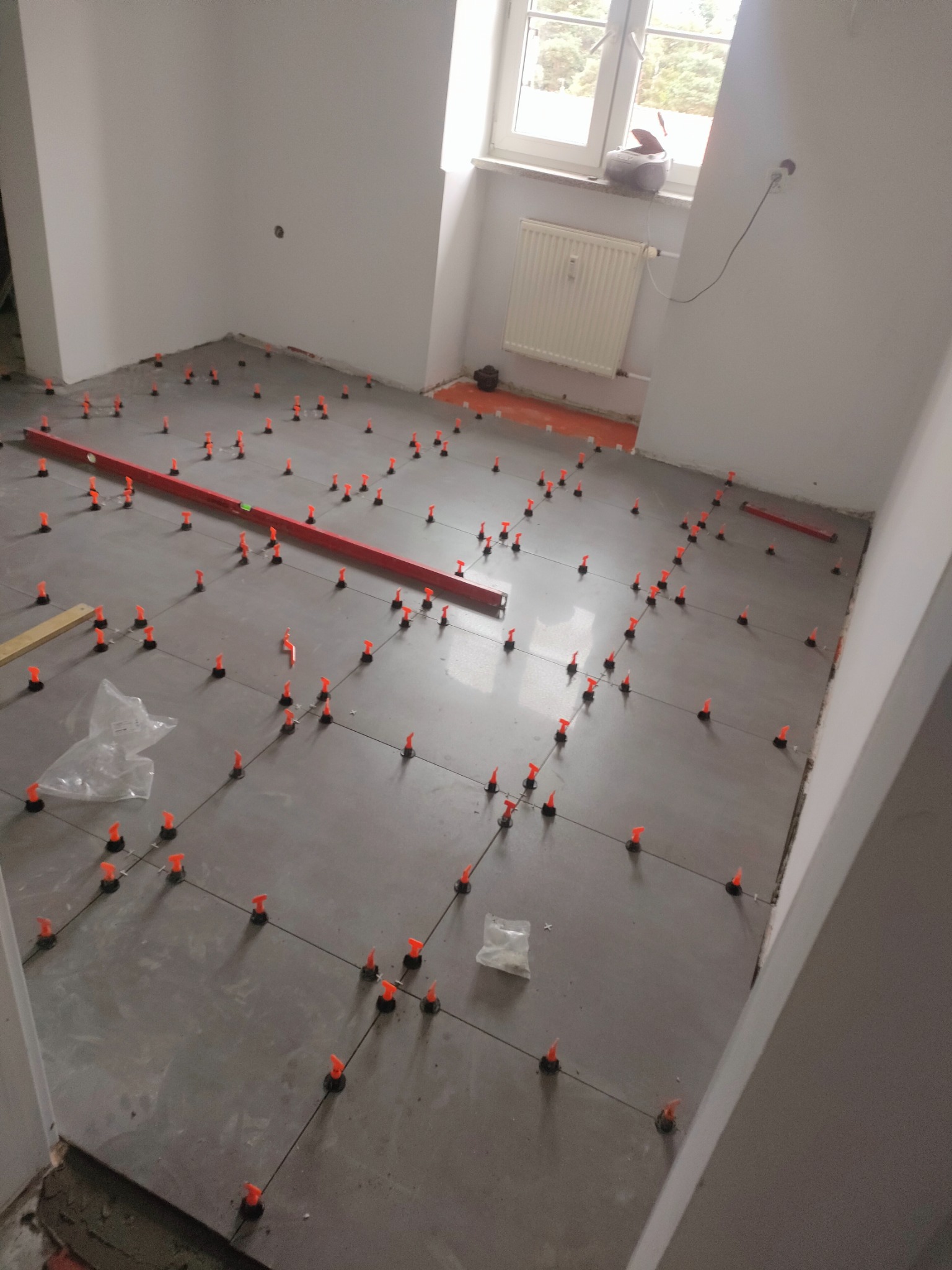 Szare płytki podłogowe w trakcie układania, z pomarańczowymi klipsami poziomującymi i poziomicą w tle, widoczne elementy grzejnika i okno.