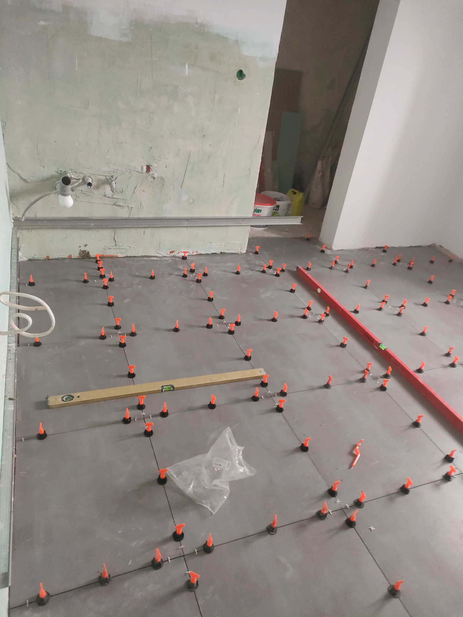 Szare płytki podłogowe w trakcie układania, z pomarańczowymi klipsami poziomującymi i dwoma poziomicami (czerwoną i drewnianą) na powierzchni, surowa ściana w tle.