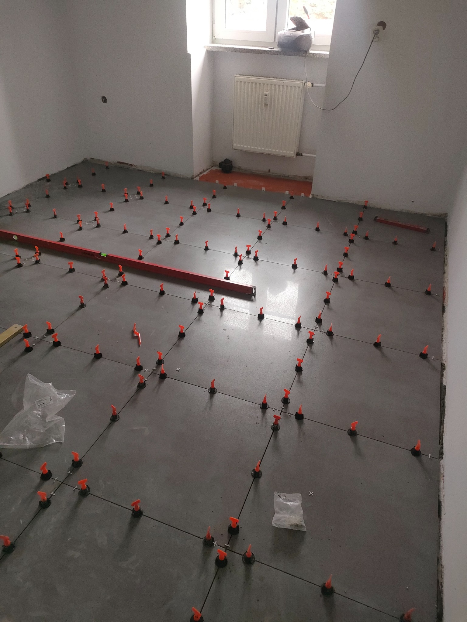 Szare płytki podłogowe w trakcie układania, z pomarańczowymi klipsami poziomującymi i krzyżykami dystansowymi, widoczna poziomica i worki z klejem.