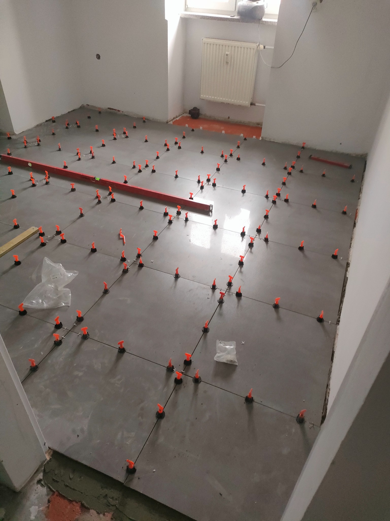 Szare płytki podłogowe w trakcie układania z użyciem pomarańczowych klipsów poziomujących, widoczna poziomica i resztki zaprawy