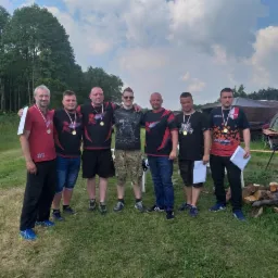 Grupa siedmiu mężczyzn z medalami na szyjach pozuje na tle zieleni, drzew i namiotu po udanym evencie sportowym lub integracyjnym.