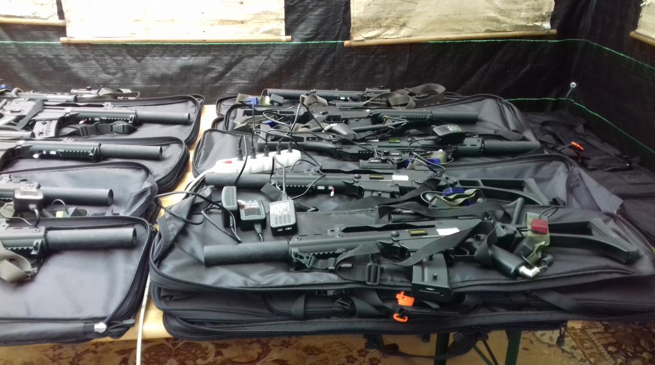 Przygotowane do transportu repliki broni laserowej, mikrofony i ładowarki w otwartych, czarnych torbach na stole z wzorzystym obrusem.