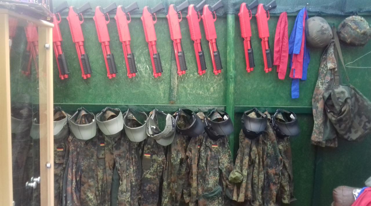 Rząd czerwonych replik broni paintballowej wiszących nad rzędem umundurowania kamuflażowego i masek ochronnych.