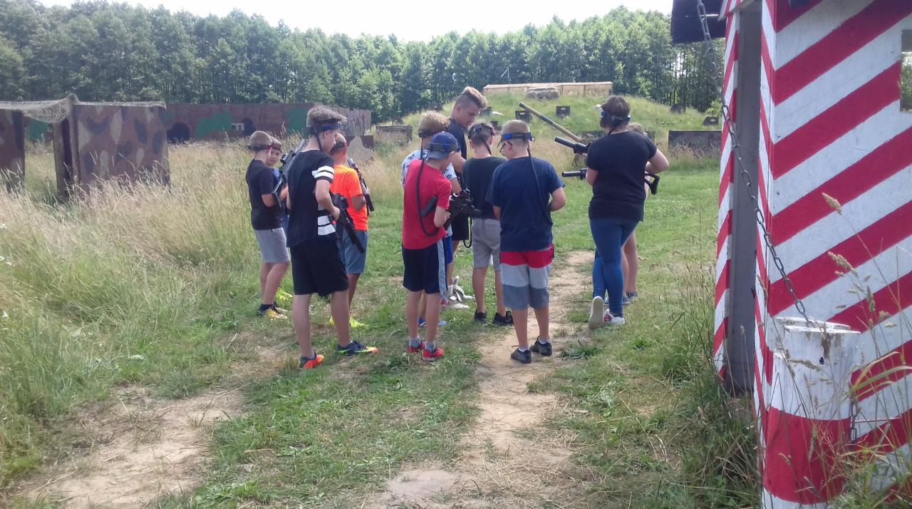 Grupa nastolatków z atrapami broni w plenerze, przygotowujących się do gry w paintball lub laser tag, na tle kamuflażowych przeszkód i drzew.