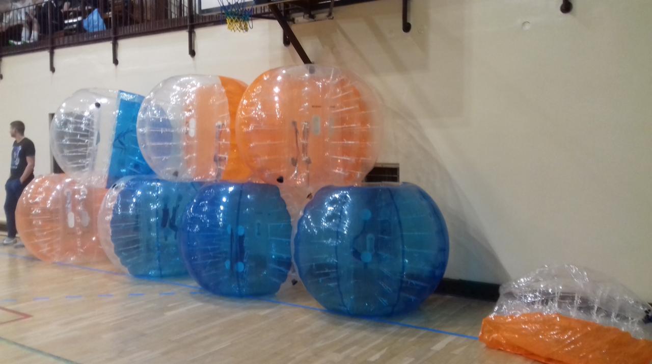 Stos kolorowych kul do bubble football w hali sportowej w Świerczach. Kule w kolorach niebieskim, pomarańczowym i transparentnym. W tle widoczny kosz do koszykówki i fragment balkonu.