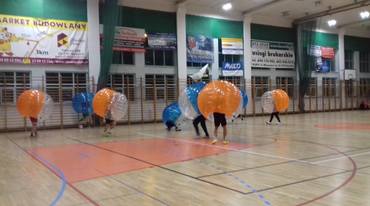 Gra Bubble Football na sali gimnastycznej: grupa osób w przezroczystych kulach z pomarańczowymi i niebieskimi elementami, widoczne reklamy na ścianach.
