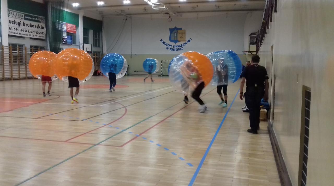 Grupa osób w przezroczystych, dmuchanych kulach (bumper balls) gra w piłkę na sali gimnastycznej, widoczny parkiet i logo szkoły na ścianie.