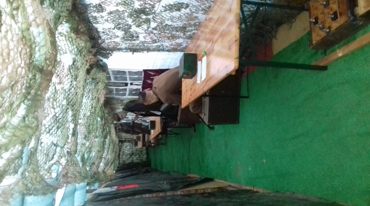 Sala przygotowana do gry terenowej z drewnianymi stołami, zieloną podłogą i siatką kamuflażową na suficie.