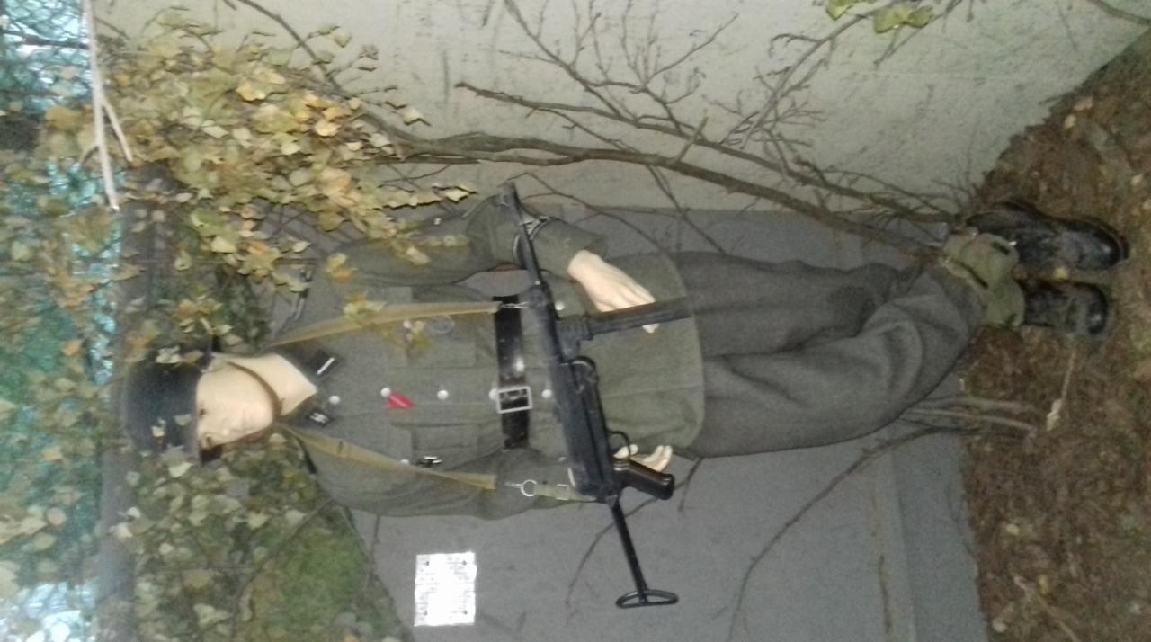 Manekin w mundurze żołnierza z karabinem MP40, ustawiony w scenerii leśnej z gałęziami i liśćmi, prawdopodobnie element ekspozycji historycznej.