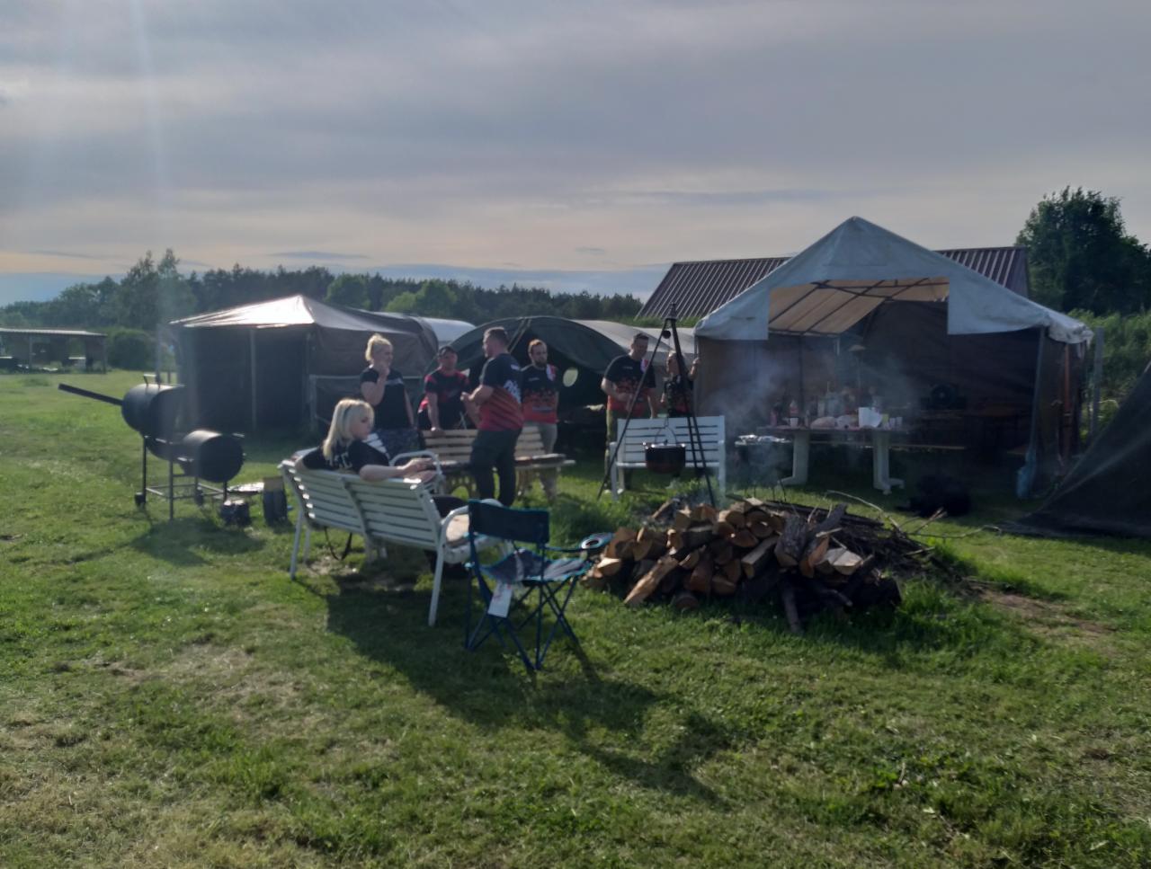 Plenerowa impreza z grillem i namiotami w tle, grupa osób w nieformalnych strojach, ognisko z drewnem na pierwszym planie.