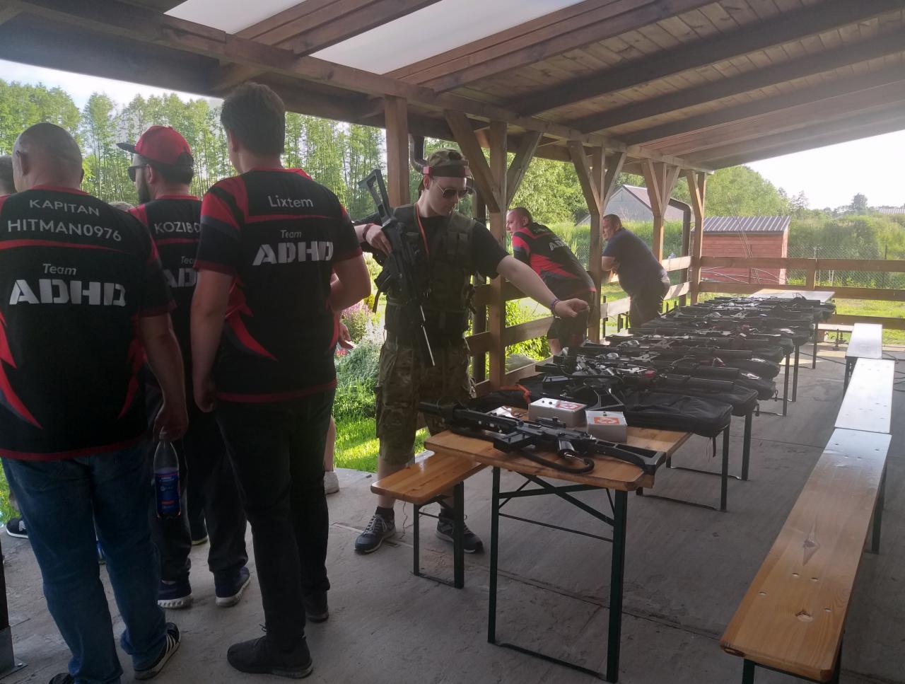 Grupa osób w koszulkach z napisem Team ADHD przygotowuje się do strzelania na strzelnicy; na stołach leżą karabiny i akcesoria strzeleckie.