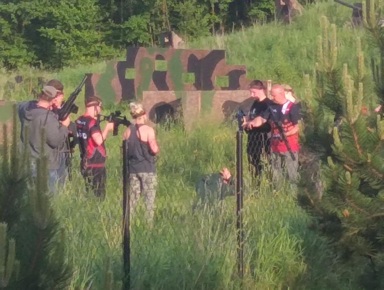 Grupa osób z replikami broni podczas rozgrywki paintballowej na zewnątrz, z kamuflażową przeszkodą w tle.