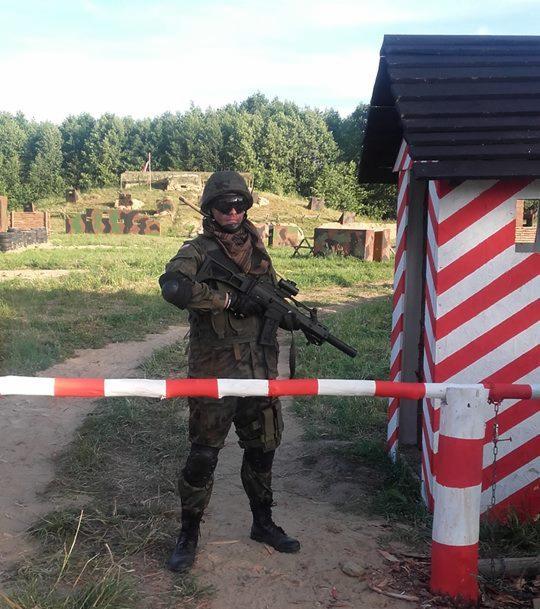 Osoba w kamuflażu, hełmie i goglach ochronnych, trzymająca replikę karabinu, stoi przed szlabanem w biało-czerwone pasy, w tle poligon z przeszkodami i budynkiem.