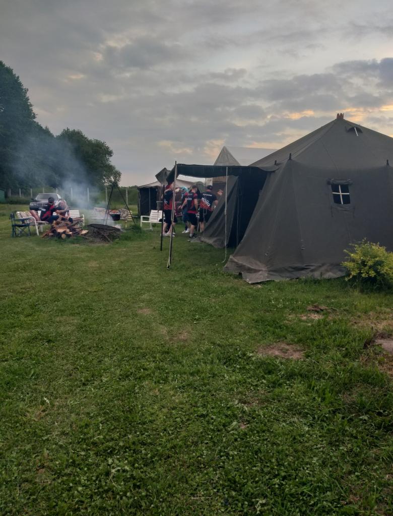 Obóz plenerowy z dużymi namiotami wojskowymi, ogniskiem i grupą osób w jednakowych koszulkach na tle zachmurzonego nieba.