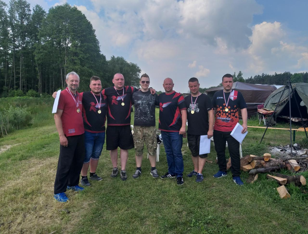 Grupa siedmiu mężczyzn z medalami na szyjach pozuje na tle zieleni, drzew i namiotu po udanym evencie sportowym lub integracyjnym.