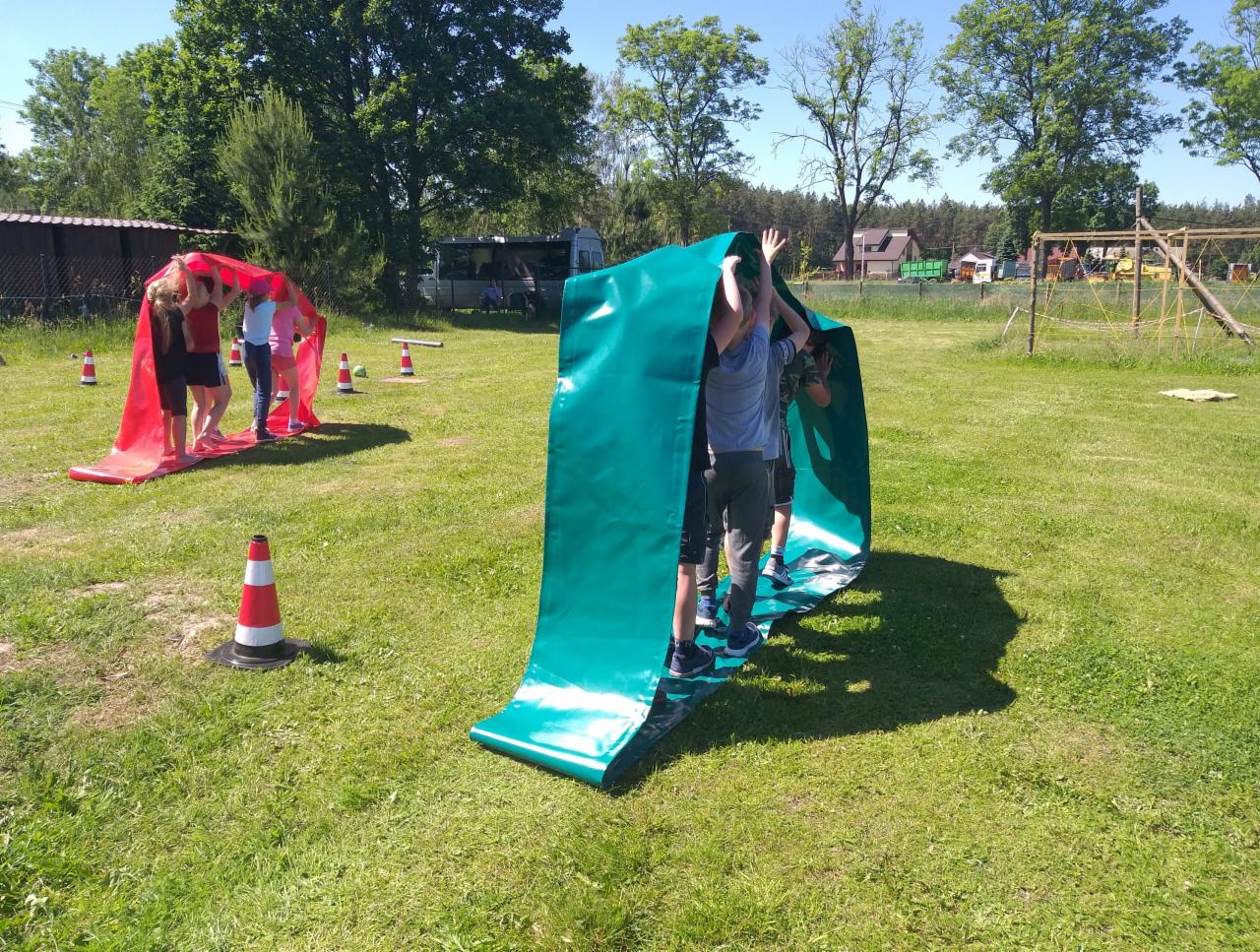 Grupa dzieci uczestniczy w zabawie z wykorzystaniem długich pasów materiału na trawie podczas imprezy plenerowej, widoczne stożki ostrzegawcze i budynek w tle.