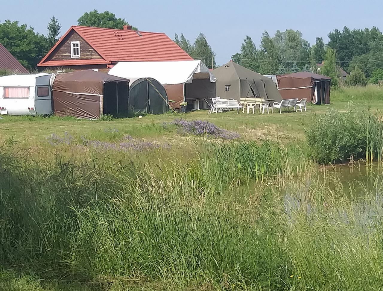 Rozległy teren z rozstawionymi namiotami, przyczepą kempingową i stołami z ławkami, tworzący plenerową przestrzeń eventową w pobliżu stawu i drewnianego domu z czerwonym dachem.