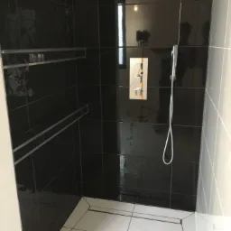 Nowoczesna kabina prysznicowa z czarnymi, błyszczącymi płytkami ściennymi, wbudowanymi półkami z metalowymi obramowaniami i białą podłogą z odpływem liniowym.