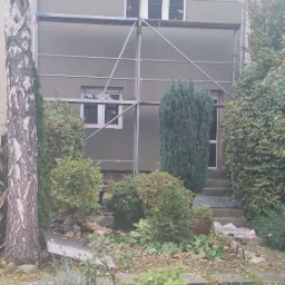 Dom jednorodzinny w trakcie tynkowania elewacji, widoczne rusztowanie, świeżo nałożony szary tynk, ogród z liśćmi jesiennymi na pierwszym planie.