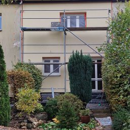 ELEWACJE Sławomir Leja - Dwupiętrowy dom w trakcie prac elewacyjnych, z rusztowaniem, oknami zabezpieczonymi folią i świeżo pomalowaną fasadą w odcieniach beżu, otoczony jesiennym ogrodem z opadłymi liśćmi.