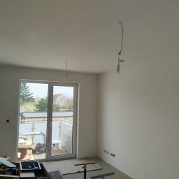 ELEWACJE Sławomir Leja - Wnętrze pokoju po świeżo wykonanych pracach wykończeniowych, widoczne gładkie, białe ściany i sufit, okno balkonowe z widokiem na zewnątrz, przewody elektryczne wystające z sufitu, resztki...