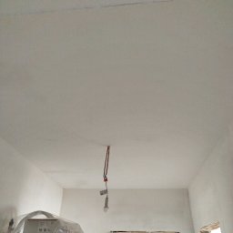 ELEWACJE Sławomir Leja - Wnętrze w trakcie remontu, ściany i sufit po nałożeniu gładzi, zabezpieczone okno folią, widoczny sprzęt malarski pod folią i prowizoryczne oświetlenie