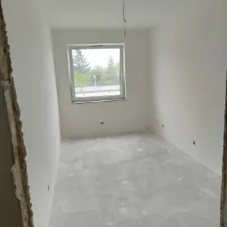 Pokój po nałożeniu pierwszej warstwy gładzi, widoczne ślady po szpachlowaniu na podłodze, wystające kable ze ściany i niedokończone obramowanie drzwi.