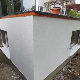 ELEWACJE Sławomir Leja - Świeżo otynkowana elewacja budynku z dwoma oknami, zabezpieczona pomarańczową folią na krawędzi dachu i doniczką na górze, widok z zewnątrz na plac budowy.