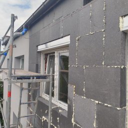 ELEWACJE Sławomir Leja - Elewacja budynku w trakcie ocieplania, widoczne płyty styropianowe grafitowe, rusztowanie i okno z roletą zewnętrzną. Szczeliny między płytami wypełnione pianką montażową.