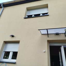 ELEWACJE Sławomir Leja - Jasna elewacja budynku z oknami z roletami, widoczny fragment rynny, daszek nad wejściem i oświetlenie zewnętrzne.