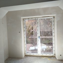 ELEWACJE Sławomir Leja - Wnętrze pokoju po szpachlowaniu ścian i sufitu, widoczne okno balkonowe z widokiem na zewnątrz, ściany przygotowane do malowania, widoczne niedoskonałości w wykończeniu.