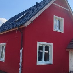 ELEWACJE Sławomir Leja - Czerwony dom jednorodzinny z czarnym dachem, białymi oknami i częściowo wykończonym wejściem, na tle błękitnego nieba i zieleni, z widoczną przenośną toaletą.