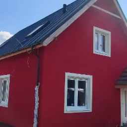 Czerwony dom jednorodzinny z czarnym dachem, białymi oknami i częściowo wykończonym wejściem, na tle błękitnego nieba i zieleni, z widoczną przenośną toaletą.