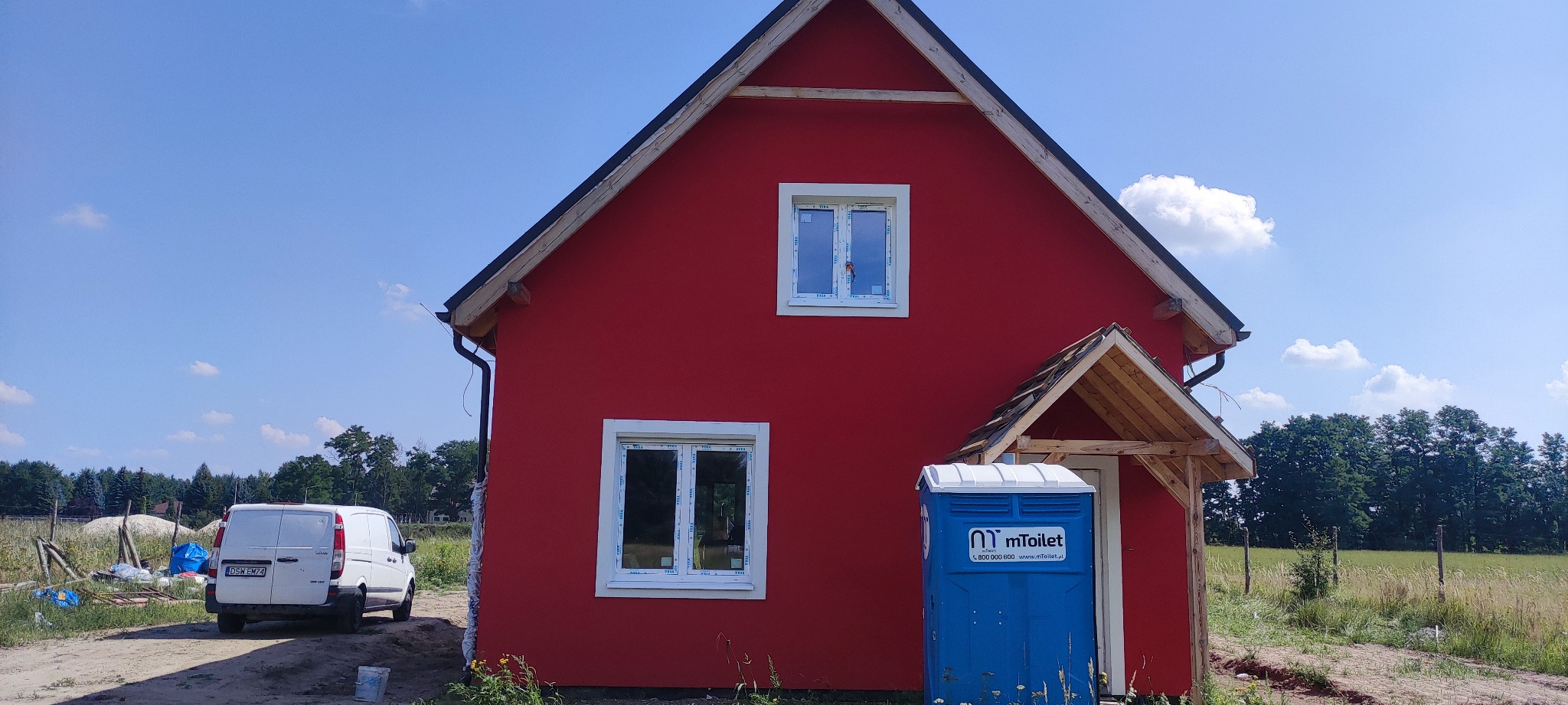 Czerwony dom jednorodzinny z białymi oknami i częściowo wykończonym zadaszeniem nad wejściem, na tle błękitnego nieba z chmurami, z toaletą przenośną i białym busem dostawczym w pobliżu.