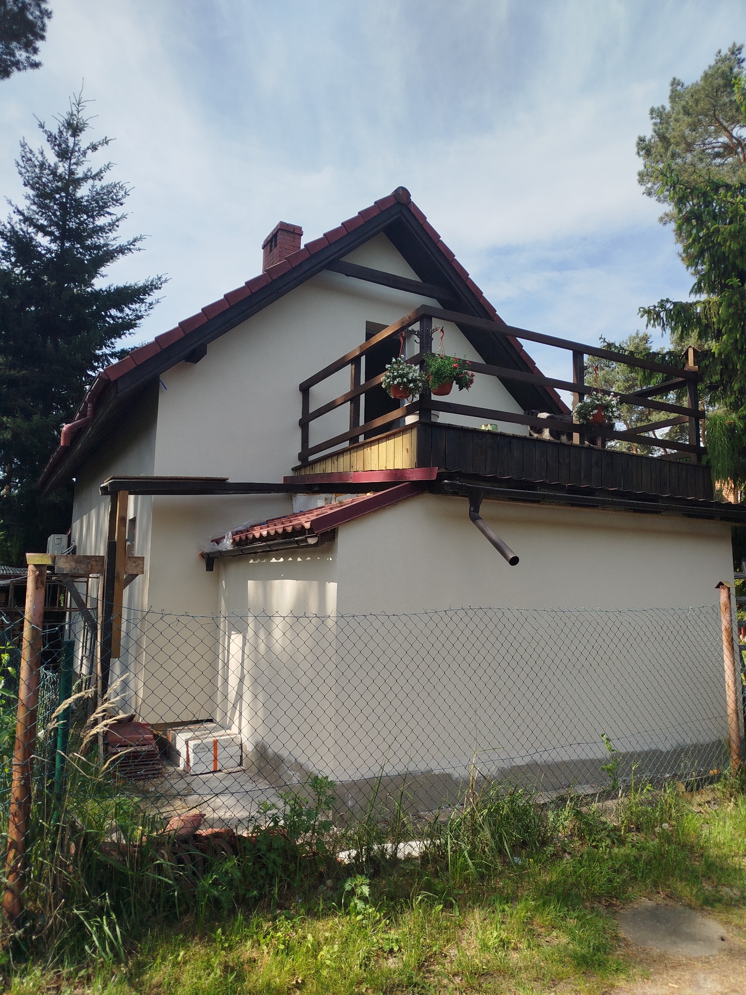 Dwupiętrowy dom z jasną elewacją, czerwonym dachem i drewnianym balkonem z kwiatami w doniczkach, widziany zza ogrodzenia z siatki.