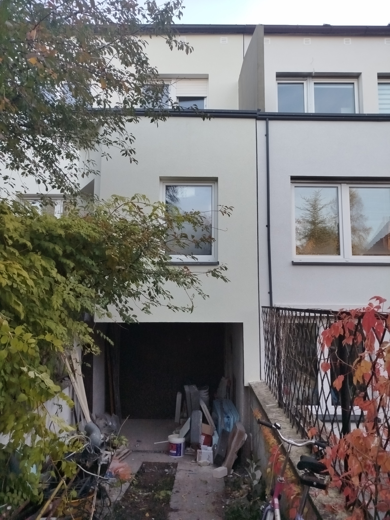 Fragment elewacji budynku po renowacji, z widocznym wejściem do garażu zagraconym materiałami budowlanymi i rowerem, zieleń częściowo zasłania fasadę.