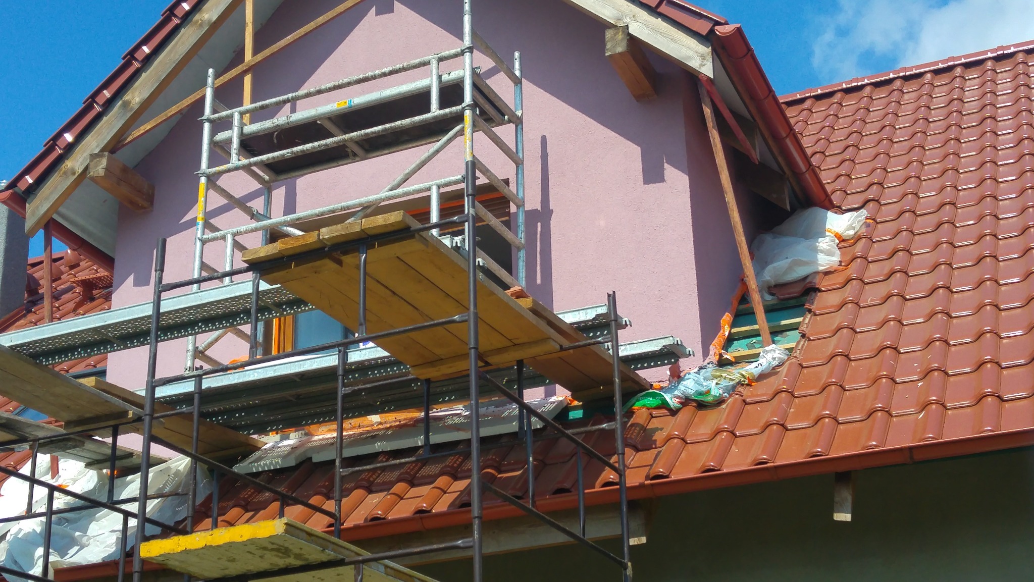 Ocieplanie poddasza budynku z różową elewacją, widoczne rusztowanie, dachówka ceramiczna i materiały izolacyjne w foliowych workach.