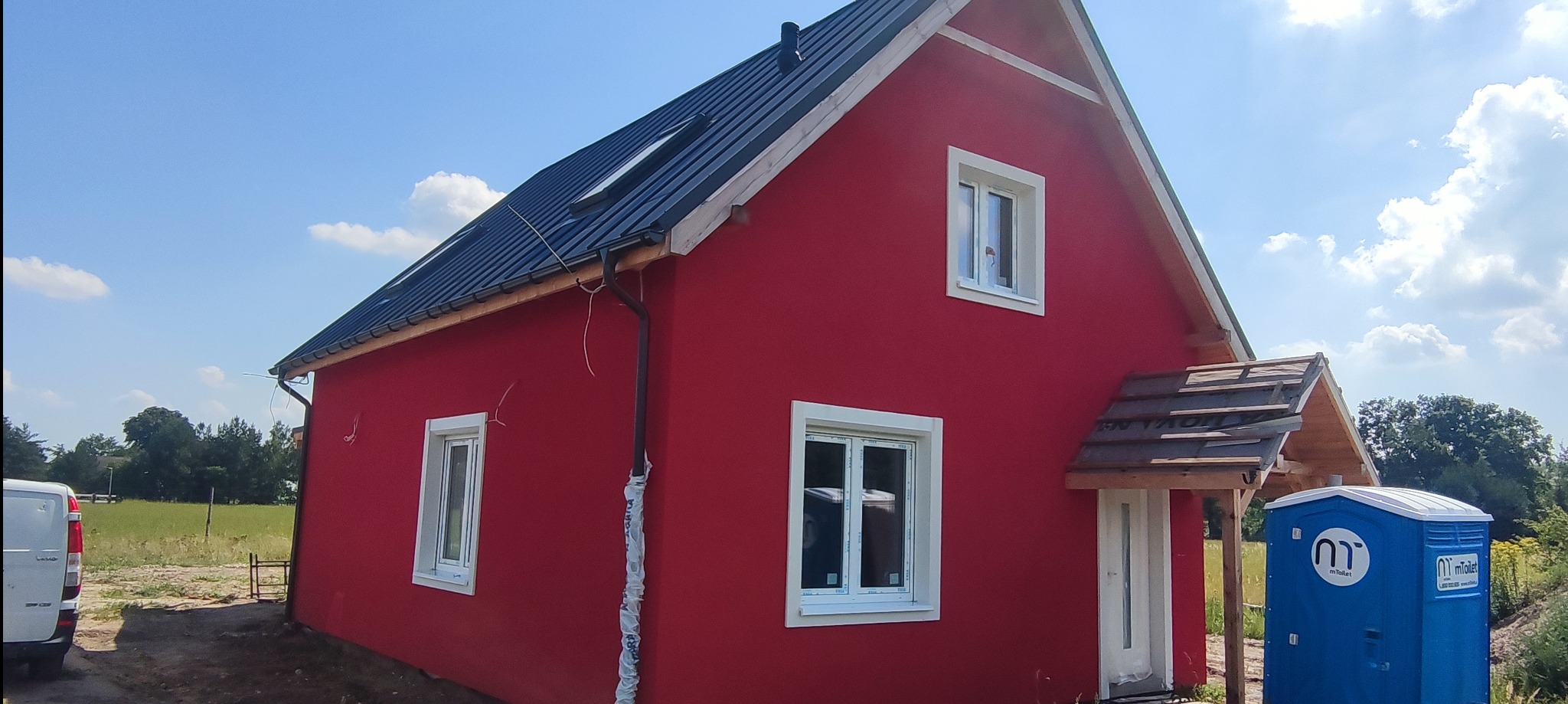 Czerwony dom jednorodzinny z czarnym dachem, białymi oknami i częściowo wykończonym wejściem, na tle błękitnego nieba i zieleni, z widoczną przenośną toaletą.