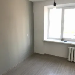 Pomalowane na szaro wnętrze pokoju z jasną podłogą, białym oknem i grzejnikiem, widoczna goła żarówka zwisająca z sufitu.
