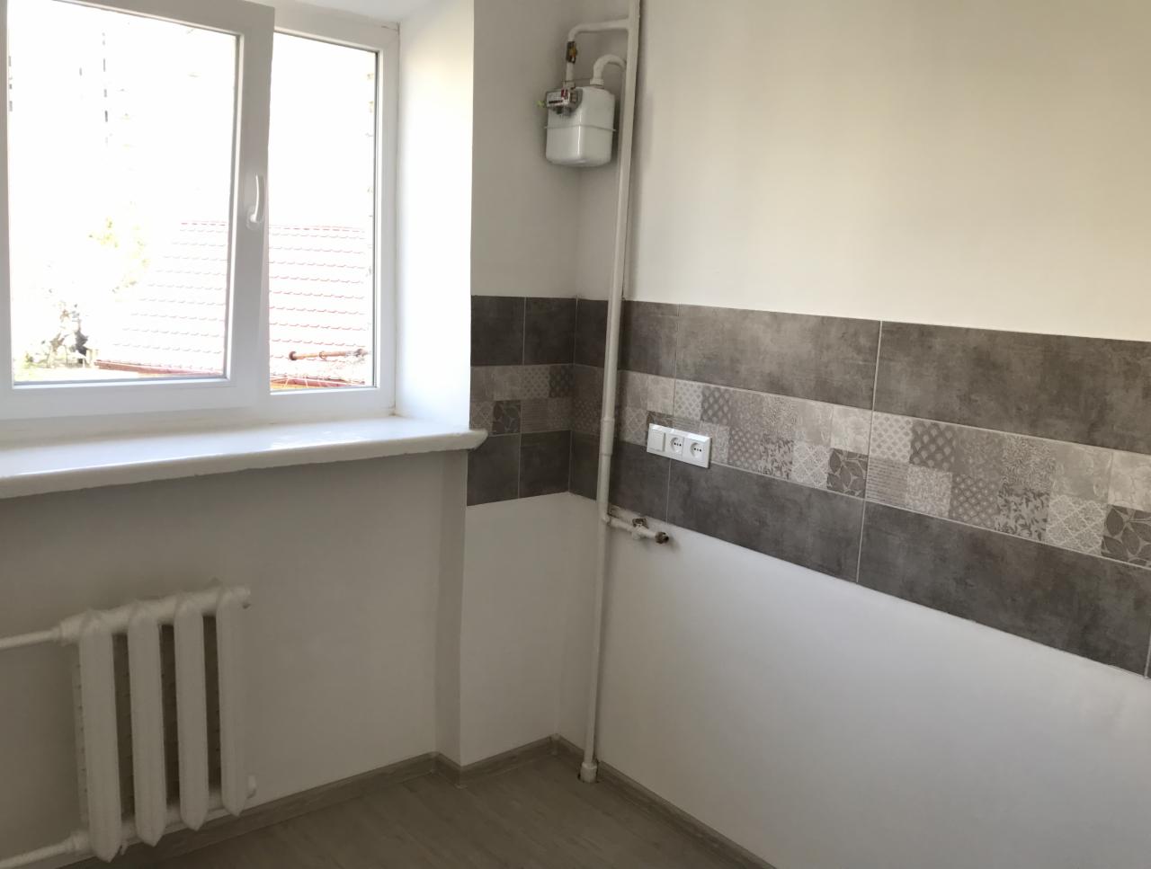 Wnętrze po odświeżeniu: ściany pomalowane na biało, dekoracyjny pas płytek w odcieniach szarości, białe okno z widokiem na zewnątrz, grzejnik przy ścianie, drewnopodobna podłoga.