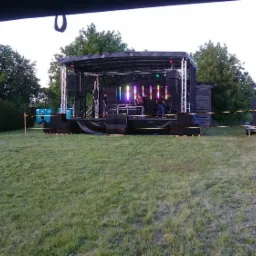 Scena plenerowa z nagłośnieniem i oświetleniem podczas koncertu, widoczni muzycy i kolorowe światła, w tle budynek i drzewa.