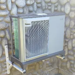 Szara zewnętrzna jednostka klimatyzacji Nibe zamontowana na białym wsporniku na ścianie z kamienia polnego.