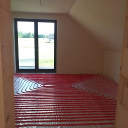 Hydro-Net - Czerwone rurki ogrzewania podłogowego ułożone spiralnie na folii izolacyjnej w surowym wnętrzu z oknem na krajobraz.