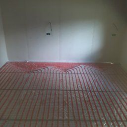 Hydro-Net - Układanie czerwonych rur systemu ogrzewania podłogowego Roth na folii izolacyjnej w pomieszczeniu z surowymi ścianami z płyt gipsowo-kartonowych, widoczne wystające kable elektryczne.