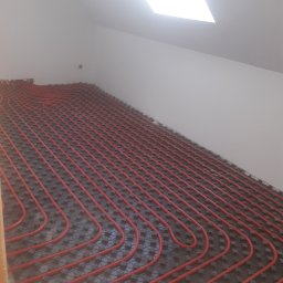 Hydro-Net - Rozłożona instalacja ogrzewania podłogowego z czerwonych rur na czarnej macie w pomieszczeniu ze skosem i oknem dachowym.