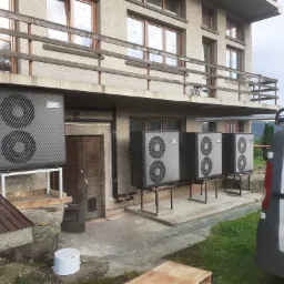 Cztery pompy ciepła zamontowane na zewnątrz budynku mieszkalnego, widoczny fragment białego samochodu dostawczego i elementy instalacyjne.