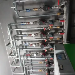 Szereg równoległych, srebrnych rur instalacji z manometrami i pompami obiegowymi, zamontowanych na szarej ścianie, z widocznym fragmentem kotła grzewczego z zieloną obudową.