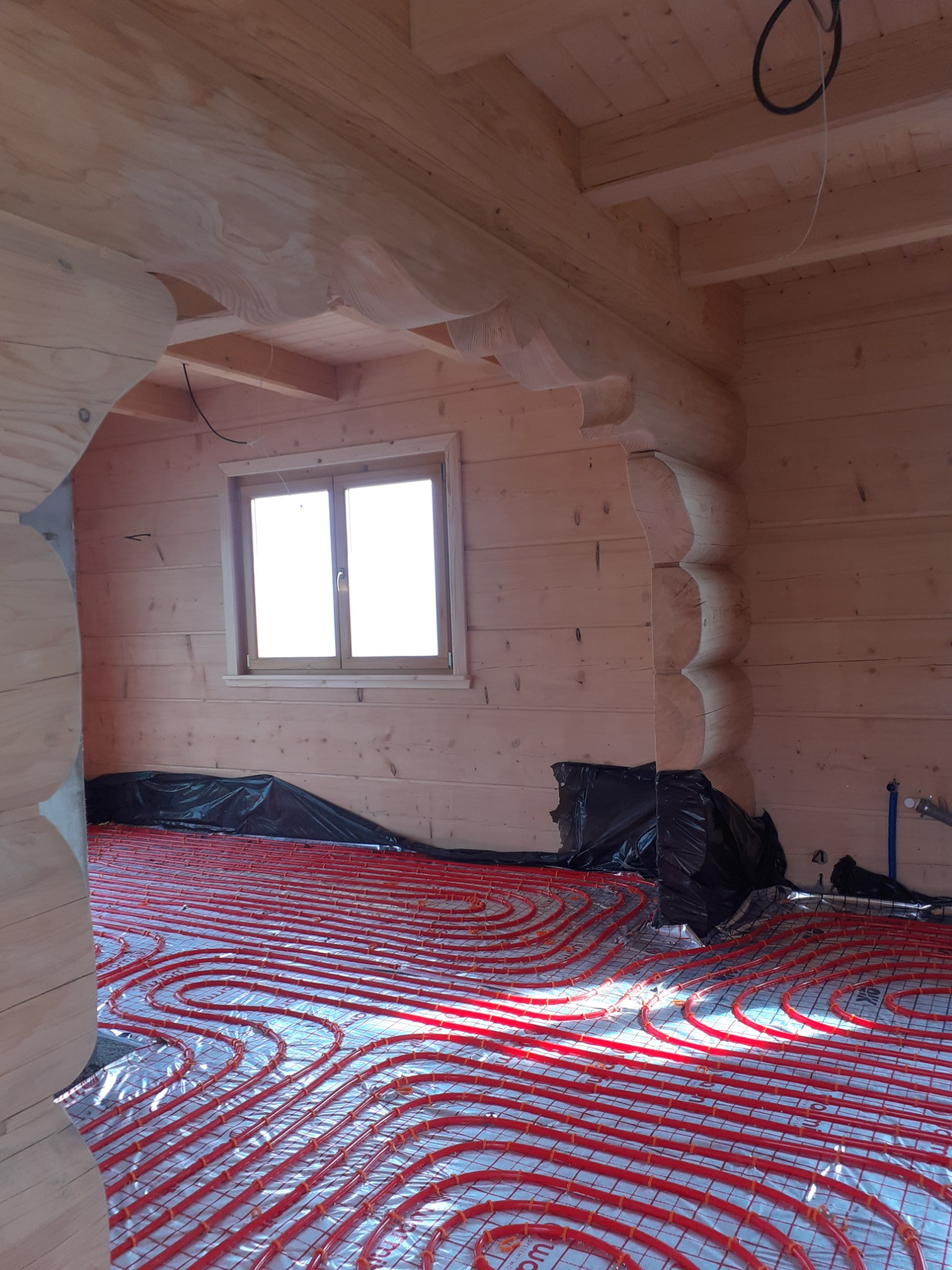 Wnętrze drewnianego domu w trakcie instalacji ogrzewania podłogowego z czerwonymi rurkami na srebrnej folii izolacyjnej, widoczne okno i elementy konstrukcyjne z bali.