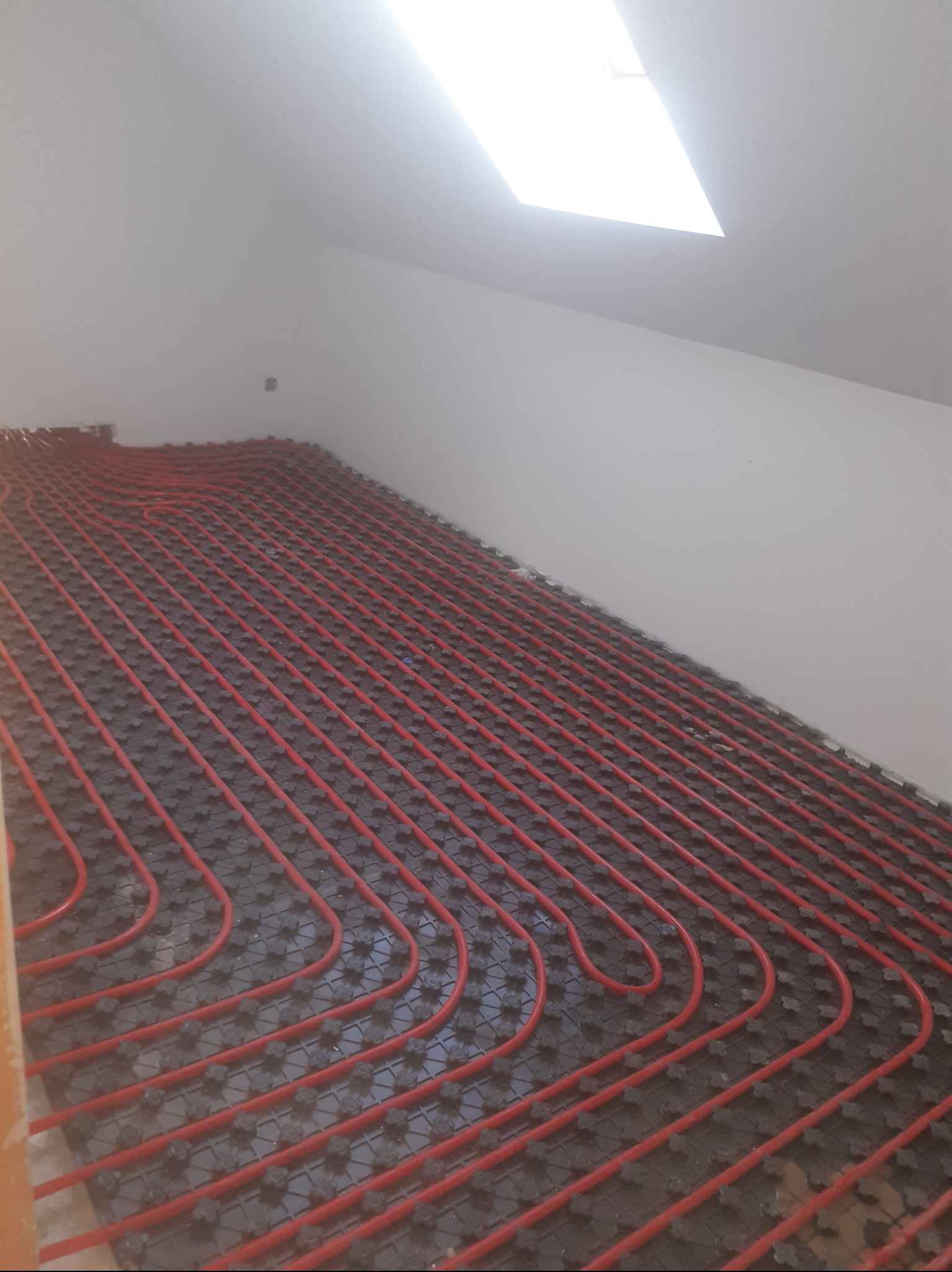 Rozłożona instalacja ogrzewania podłogowego z czerwonych rur na czarnej macie w pomieszczeniu ze skosem i oknem dachowym.