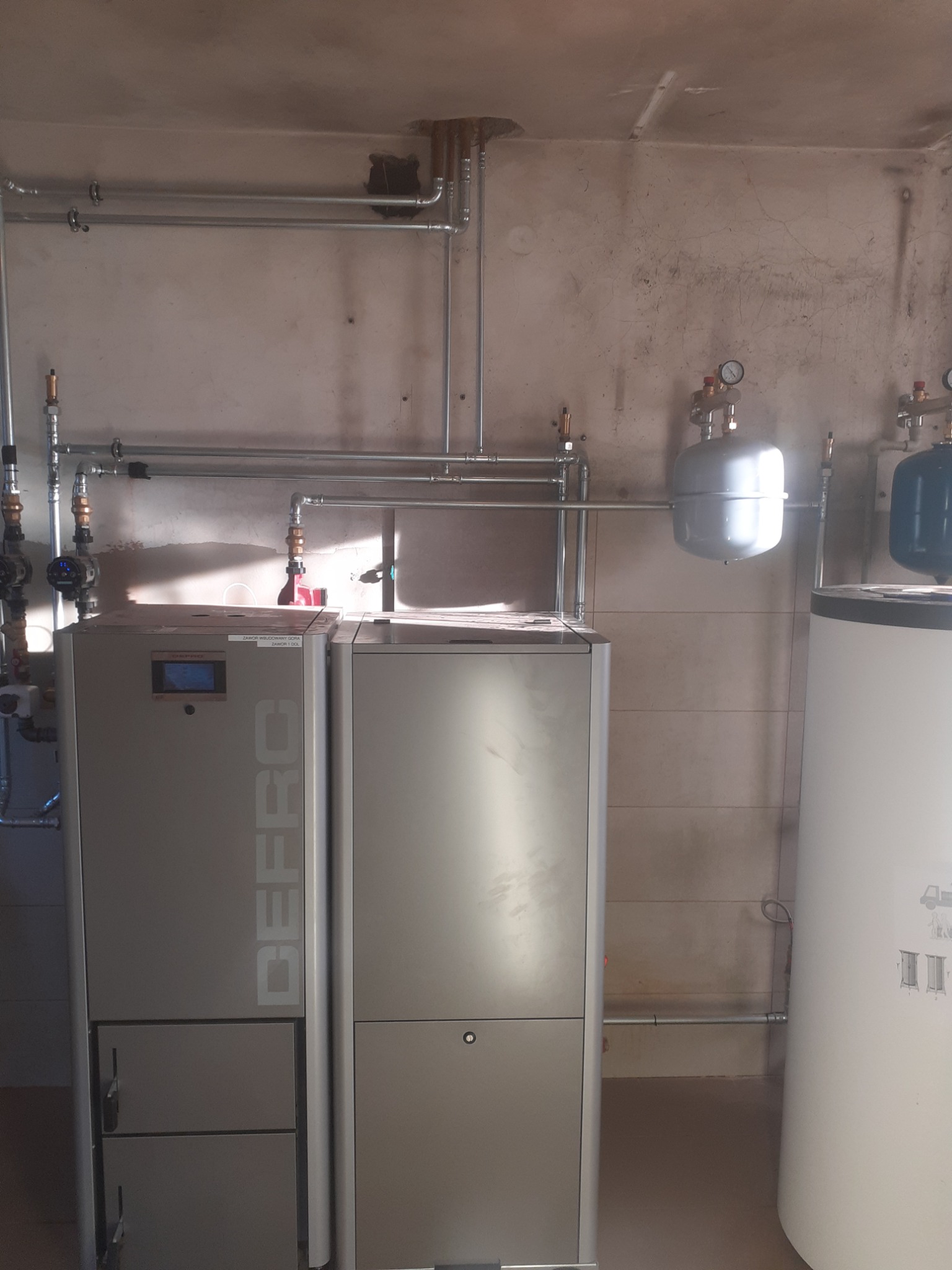 Szara kotłownia z dwoma piecami, zbiornikiem na wodę i instalacją hydrauliczną na tle surowej ściany.